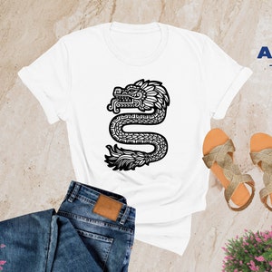 May include: White t-shirt with a black and white graphic of a stylized serpent with a feathered headdress. The serpent is facing left and has a detailed design. The t-shirt is on a light brown surface with a pair of sandals and a pair of jeans.
