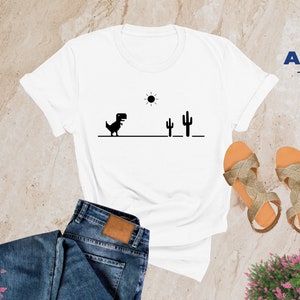 May include: White t-shirt featuring a black dinosaur graphic, sun, and cacti. The shirt is paired with blue jeans and gold sandals. The word "WHITE" is in a white box in the upper left corner. The Alpine Design logo is in the upper right corner.