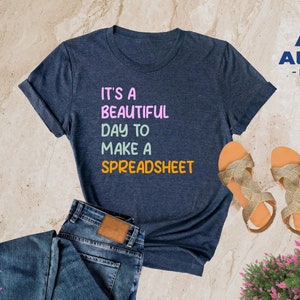 May include: Heather navy blue t-shirt with the text "It's a beautiful day to make a spreadsheet" in colorful letters. The shirt is laid out on a light brown surface with a pair of gold sandals and blue jeans.