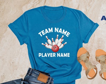 May include: A heather deep teal t-shirt with a bowling ball and pins graphic. The text "TEAM NAME" is above the graphic and "PLAYER NAME" is below the graphic. The t-shirt is on a light brown surface with a pair of gold sandals and a pair of blue jeans.