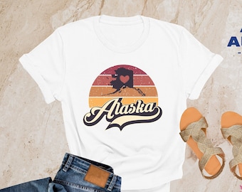 May include: White t-shirt with a vintage-style graphic of the state of Alaska with a red heart in the middle. The graphic is set against a sunset background with the word "Alaska" in a retro font.