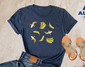 May include: A heather navy blue t-shirt with a graphic of yellow bananas. The t-shirt is on a light brown surface with a pair of gold sandals and blue jeans. The text "ALPINE -DESIGN- " is on the t-shirt.
