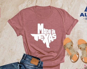May include: A heather mauve t-shirt with the text "Made in Texas" in white, printed in the shape of the state of Texas. The shirt is laid out on a light brown surface with a pair of gold sandals and a pair of blue jeans.