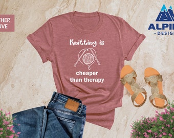 May include: Heather mauve t-shirt with the text "Knitting is cheaper than therapy" and a white outline of yarn and knitting needles in a heart shape. The t-shirt is laid out on a light brown textured surface with a pair of gold sandals and a pair of blue jeans.