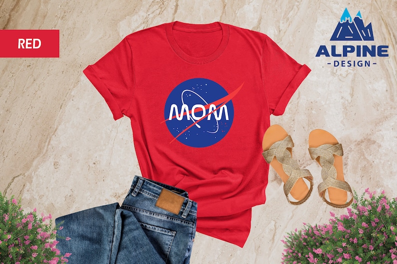 May include: Red t-shirt with a blue and white NASA logo that says "MOM" on it. The shirt is laid out on a light brown surface with a pair of gold sandals and blue jeans.