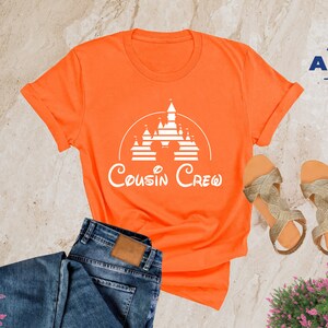 Pode incluir: Camiseta laranja com um gr&aacute;fico branco de um castelo e o texto "Cousin Crew".
