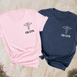 May include: Two t-shirts, one pink and one heather navy, with a medical caduceus symbol and the text "OB-GYN" printed on the front.