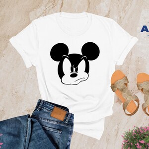 May include: White t-shirt with a black and white image of Mickey Mouse with an angry expression. The shirt is on a light brown surface with a pair of gold sandals and a pair of blue jeans.