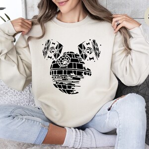 May include: A sand-colored sweatshirt with a black graphic of the Death Star from Star Wars, with two TIE fighters on either side of it. The graphic is designed to look like Mickey Mouse ears.