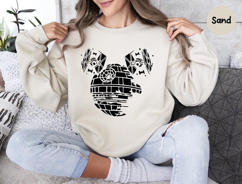 May include: A sand-colored sweatshirt with a black graphic of the Death Star from Star Wars, with two TIE fighters on either side of it. The graphic is designed to look like Mickey Mouse ears.