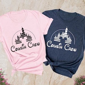 Pode incluir: Duas camisetas com um gr&aacute;fico preto e branco de um castelo com o texto "Cousin Crew" em uma fonte de escrita cursiva preto e branco. As camisetas s&atilde;o rosa e azul marinho mesclado.