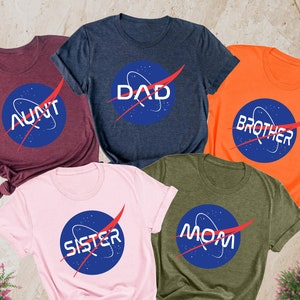May include: Five t-shirts with a space-themed design featuring the words "Aunt", "Dad", "Brother", "Sister", and "Mom" in a blue circle with a red swoosh. The shirts are in various colors: maroon, navy blue, orange, pink, and olive green.