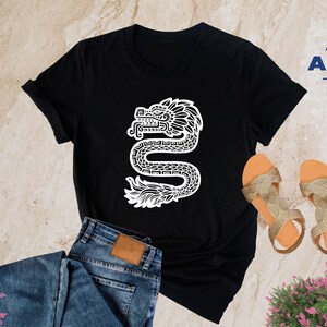 Może przedstawiać: Czarny T-shirt z białym nadrukiem stylizowanej węża z pi&oacute;ropuszem. Tekst "ALPINE -DESIGN- " znajduje się w prawym g&oacute;rnym rogu obrazu.