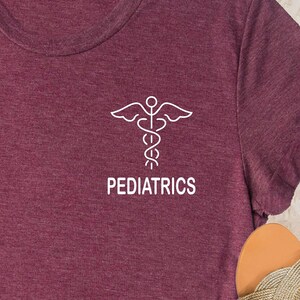 May include: Burgundy t-shirt featuring a white medical symbol and the word "PEDIATRICS" in a clean, sans-serif font. The shirt has a crew neck and a soft, textured appearance. The design is centered on the chest.