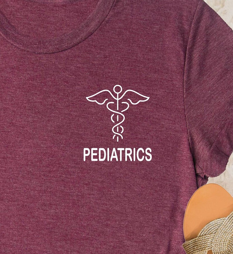 May include: Burgundy t-shirt featuring a white medical symbol and the word "PEDIATRICS" in a clean, sans-serif font. The shirt has a crew neck and a soft, textured appearance. The design is centered on the chest.