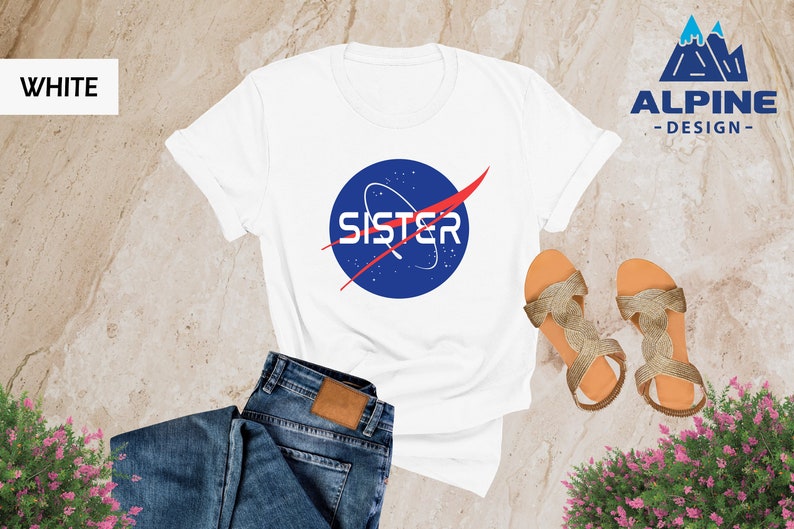 May include: White t-shirt with a blue and red NASA-inspired logo that says "SISTER" on it. The shirt is being modeled with blue jeans and gold sandals.