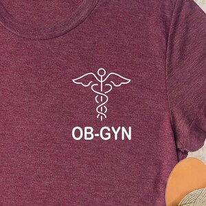 May include: A maroon t-shirt with a white outline of a medical caduceus symbol and the text "OB-GYN".