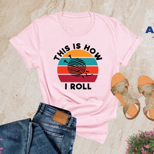 May include: Pink t-shirt with a retro sunset graphic and the text "This is how I roll" with a ball of yarn and knitting needles in the center of the graphic.