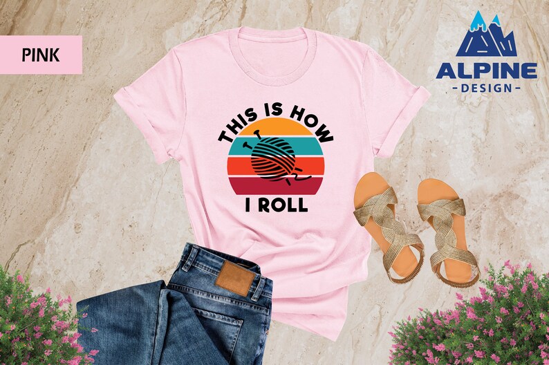 May include: Pink t-shirt with a retro sunset graphic and the text "This is how I roll" with a ball of yarn and knitting needles in the center of the graphic.