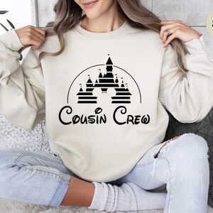 Pode incluir: Uma camisa de moletom na cor areia com um gr&aacute;fico preto de um castelo e o texto "Cousin Crew" em uma fonte inspirada na Disney.