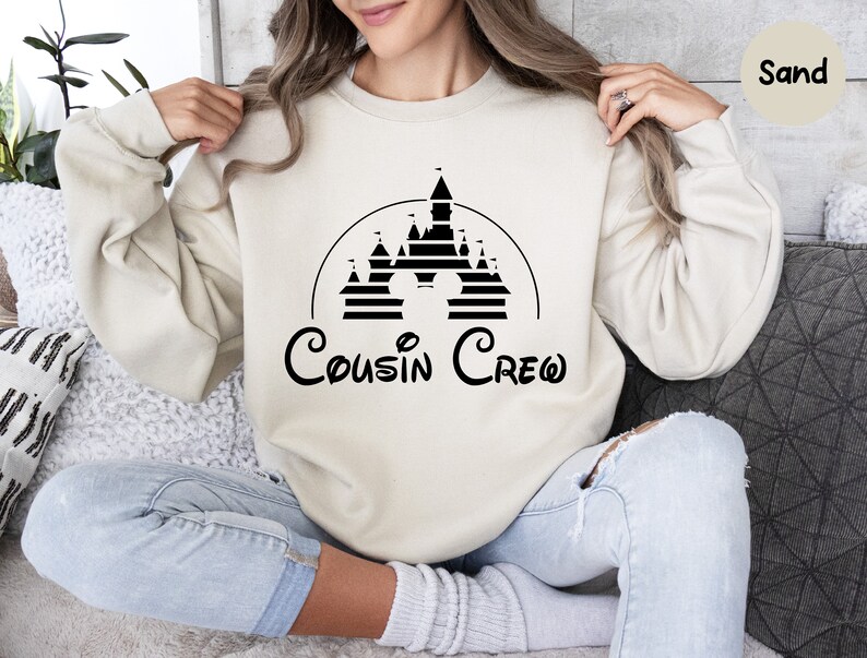 Pode incluir: Uma camisa de moletom na cor areia com um gr&aacute;fico preto de um castelo e o texto "Cousin Crew" em uma fonte inspirada na Disney.