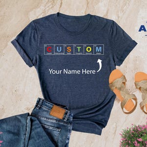 May include: Heather navy blue t-shirt with a graphic of the word "CUSTOM" spelled out using chemical element symbols. The text "Your Name Here" is printed below the graphic with an arrow pointing to it. The t-shirt is displayed on a light brown surface with a pair of gold sandals and blue jeans.