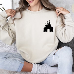 Op de afbeelding: Een zandkleurig sweatshirt met een zwart silhouet van een kasteel op de voorkant. Het sweatshirt wordt gedragen door een persoon die op een bank zit.