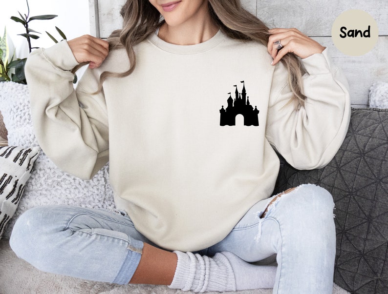 Op de afbeelding: Een zandkleurig sweatshirt met een zwart silhouet van een kasteel op de voorkant. Het sweatshirt wordt gedragen door een persoon die op een bank zit.