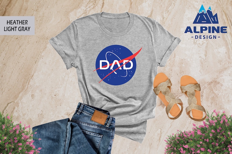 May include: A heather light gray t-shirt with a blue and red NASA-inspired logo that says "DAD" on it. The shirt is laid out on a light brown surface with a pair of gold sandals and blue jeans.