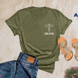 May include: A heather olive t-shirt with a white outline of a medical caduceus symbol and the text "OB-GYN".