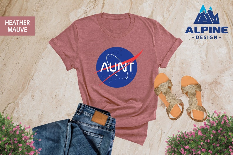 May include: Heather mauve t-shirt with a blue and red NASA-inspired graphic that says "AUNT". The shirt is laid out on a light brown surface with a pair of gold sandals and blue jeans.