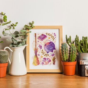 Puede incluir: Una impresi&oacute;n enmarcada con una ilustraci&oacute;n caprichosa de un caldero, una escoba, un libro y hongos en tonos de morado, amarillo y verde. La impresi&oacute;n est&aacute; rodeada de plantas en macetas, incluyendo suculentas y eucalipto.