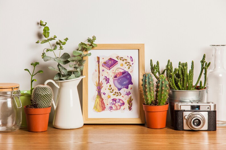 Puede incluir: Una impresi&oacute;n enmarcada con una ilustraci&oacute;n caprichosa de un caldero, una escoba, un libro y hongos en tonos de morado, amarillo y verde. La impresi&oacute;n est&aacute; rodeada de plantas en macetas, incluyendo suculentas y eucalipto.