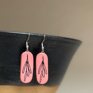 May include: A pair of pink ceramic earrings with a black floral design. The earrings are oval shaped and have silver hooks.