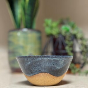 May include: A small, handmade ceramic bowl with a dark blue glaze and a gold rim. The bowl is sitting on a light brown surface.