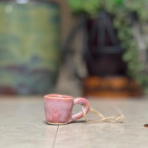 May include: A small, handmade ceramic mug in shades of pink and red, with a looped handle. A natural twine is attached to the handle. The mug is sitting on a light-colored surface, with blurred greenery in the background.