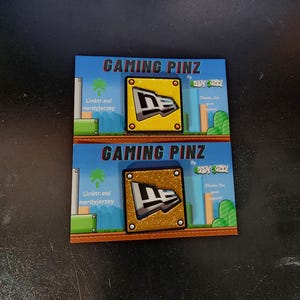 May include: Two cards featuring a gold and silver New Era logo pin on each. The cards are blue and green with the text "Gaming Pinz" and "Nerdy Jersey" on them.
