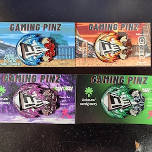 May include: Four colorful enamel pins featuring a stylized "DE" logo with different backgrounds. The pins are displayed on cards with the text "Gaming Pinz" and a link to a website.