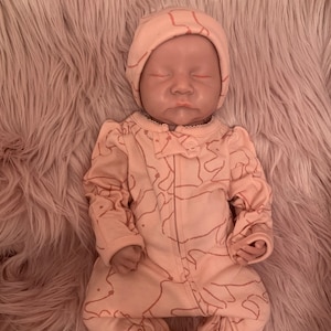 May include: A pink baby onesie with a pink and white hat. The onesie has a pattern of abstract shapes and the hat has a simple design. The baby is sleeping peacefully.