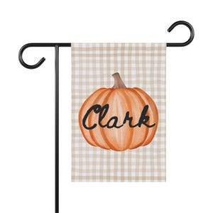 Puede incluir: Una bandera de jardín con un diseño de calabaza naranja y el nombre "Clark" en escritura negra. La bandera tiene un fondo a cuadros beige y blanco y se muestra en un soporte de metal negro.