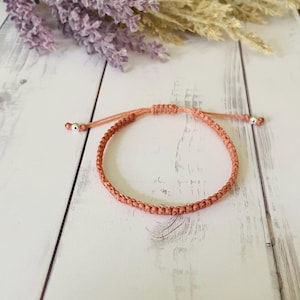 May include: A peach-colored cord bracelet featuring a braided design with an adjustable sliding knot closure. The ends of the cord are finished with small, polished silver-toned metal beads, providing a secure and flexible fit for the wrist.