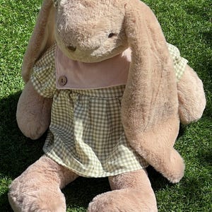 May include: A plush brown bunny wearing a green and white gingham dress with a pink bib and a bow on its head. The bunny is sitting on green grass.
