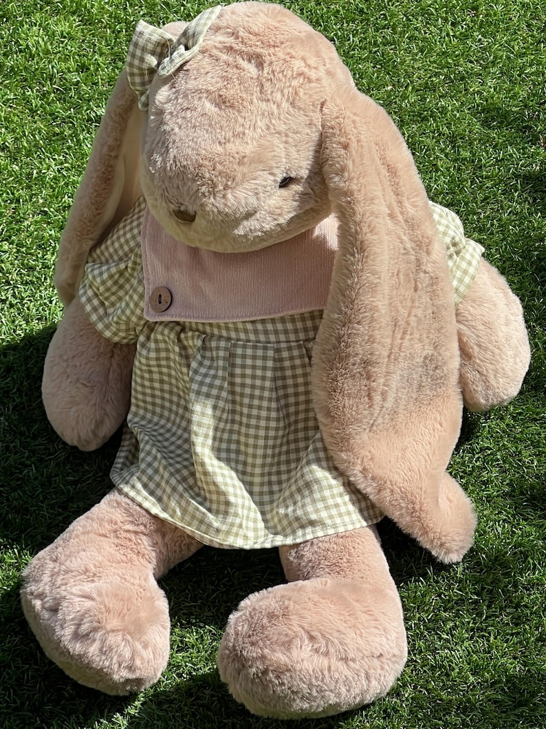 May include: A plush brown bunny wearing a green and white gingham dress with a pink bib and a bow on its head. The bunny is sitting on green grass.