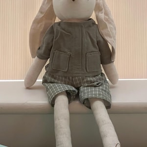May include: A soft toy bunny with long ears, dressed in a green jacket and plaid shorts. The bunny has a pale face and limbs, with brown eyes and a brown nose. It is wearing brown shoes with laces. The doll is sitting on a white surface.