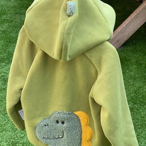 May include: A light green fleece jacket featuring a dinosaur design on the back. The dinosaur has a textured, light green face and a yellow, textured spine. The hood is adorned with three pom-poms.