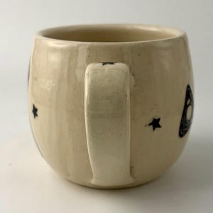 K&ouml;nnte beinhalten: Ein cremefarbener Keramikbecher mit rundem K&ouml;rper und einem stabilen Henkel. Die Au&szlig;enfl&auml;che ist mit kleinen, handgemalten schwarzen Sternen und einem dunklen, wirbelnden grafischen Element verziert. Die Glasur hat ein glattes, gl&auml;nzendes Finish.