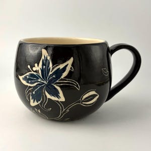 May include: A rounded ceramic mug with a glossy black glaze featuring a hand-painted lily design in shades of dark blue and cream. The mug has a curved handle on the right side and a smooth, light-colored interior finish, showcasing a floral motif.
