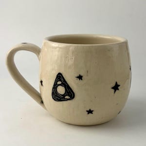 K&ouml;nnte beinhalten: Ein cremefarbener Keramikbecher mit rundem K&ouml;rper und geschwungenem Henkel. Die Au&szlig;enseite ist mit handgezeichneten schwarzen Tintenillustrationen einer dreieckigen Planchette und verschiedenen kleinen Sternen auf der gl&auml;nzenden Glasur verziert.