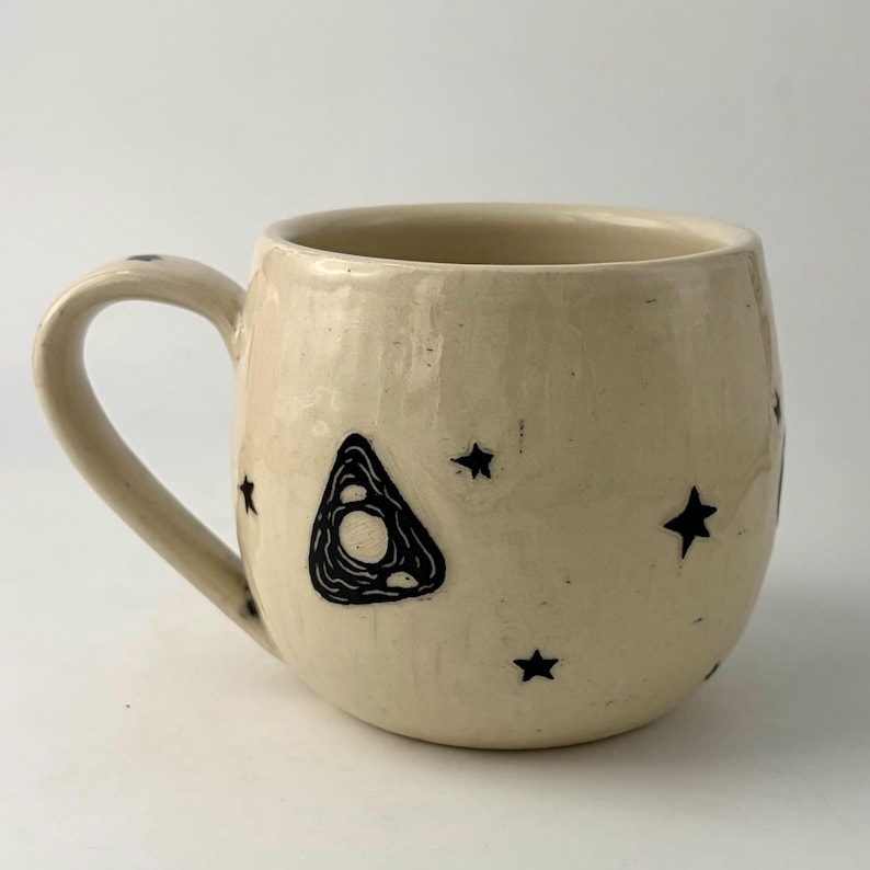 K&ouml;nnte beinhalten: Ein cremefarbener Keramikbecher mit rundem K&ouml;rper und geschwungenem Henkel. Die Au&szlig;enseite ist mit handgezeichneten schwarzen Tintenillustrationen einer dreieckigen Planchette und verschiedenen kleinen Sternen auf der gl&auml;nzenden Glasur verziert.