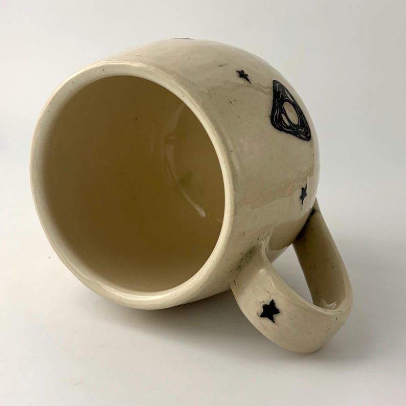 K&ouml;nnte beinhalten: Ein cremefarbener Keramikbecher mit gl&auml;nzender Oberfl&auml;che, verziert mit handgemalten schwarzen Sternen und einem Planchette-Motiv auf der Au&szlig;enseite. Der Becher hat einen abgerundeten K&ouml;rper, eine weite &Ouml;ffnung und einen stabilen Henkel.